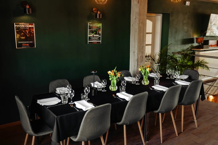 An elegant dining table set with black chairs and white place settings, fresh flowers, and glasses against a deep green wall with warm lighting.