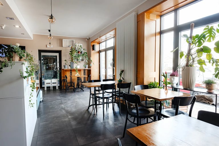 Modern cafe interior with wooden tables, black chairs, large windows, plants, and natural lighting throughout bright dining space