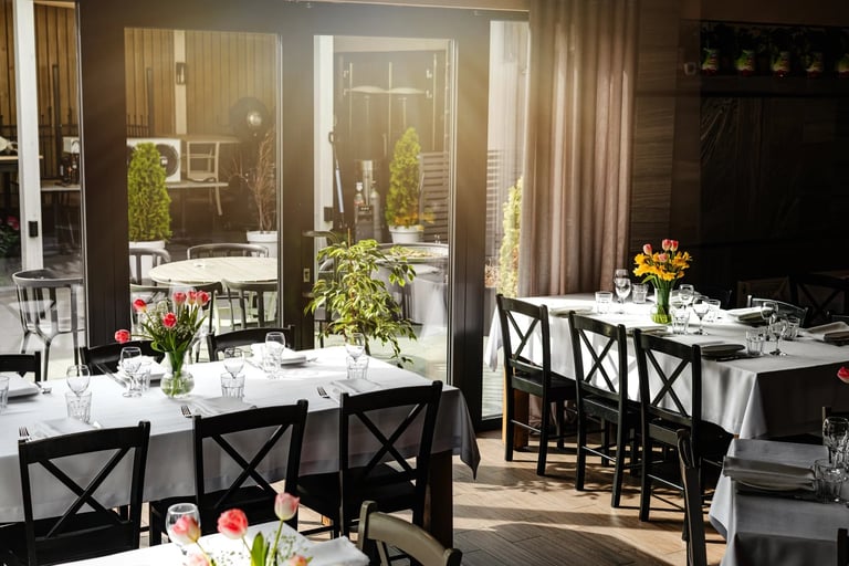 Elegant fine dining restaurant interior with white tablecloths, black X-back chairs, fresh flowers, and French doors leading to an outdoor patio.
