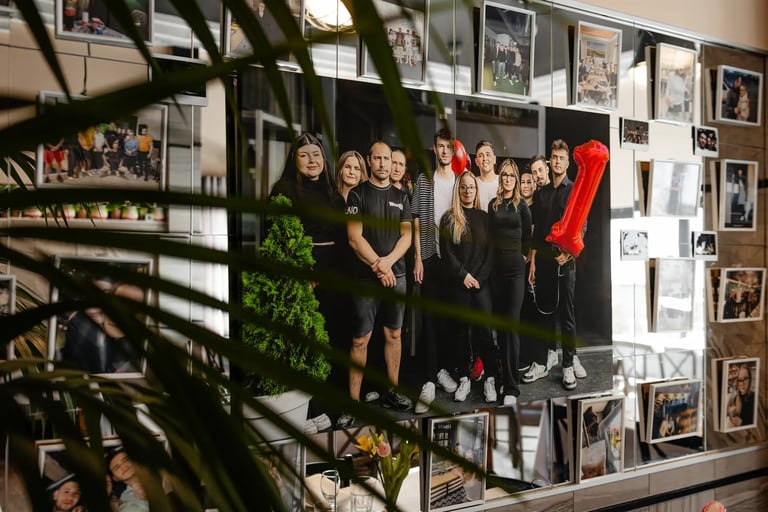 Group of people in dark clothing standing together in front of a wall covered with framed photographs and memories, with green plants in the foreground.