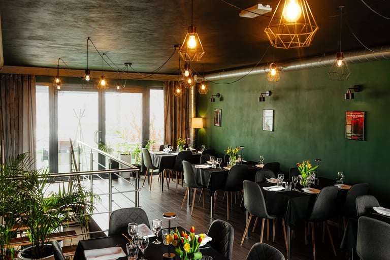 Modern restaurant interior with green accent wall, black tables, hanging pendant lights, and large windows overlooking a balcony