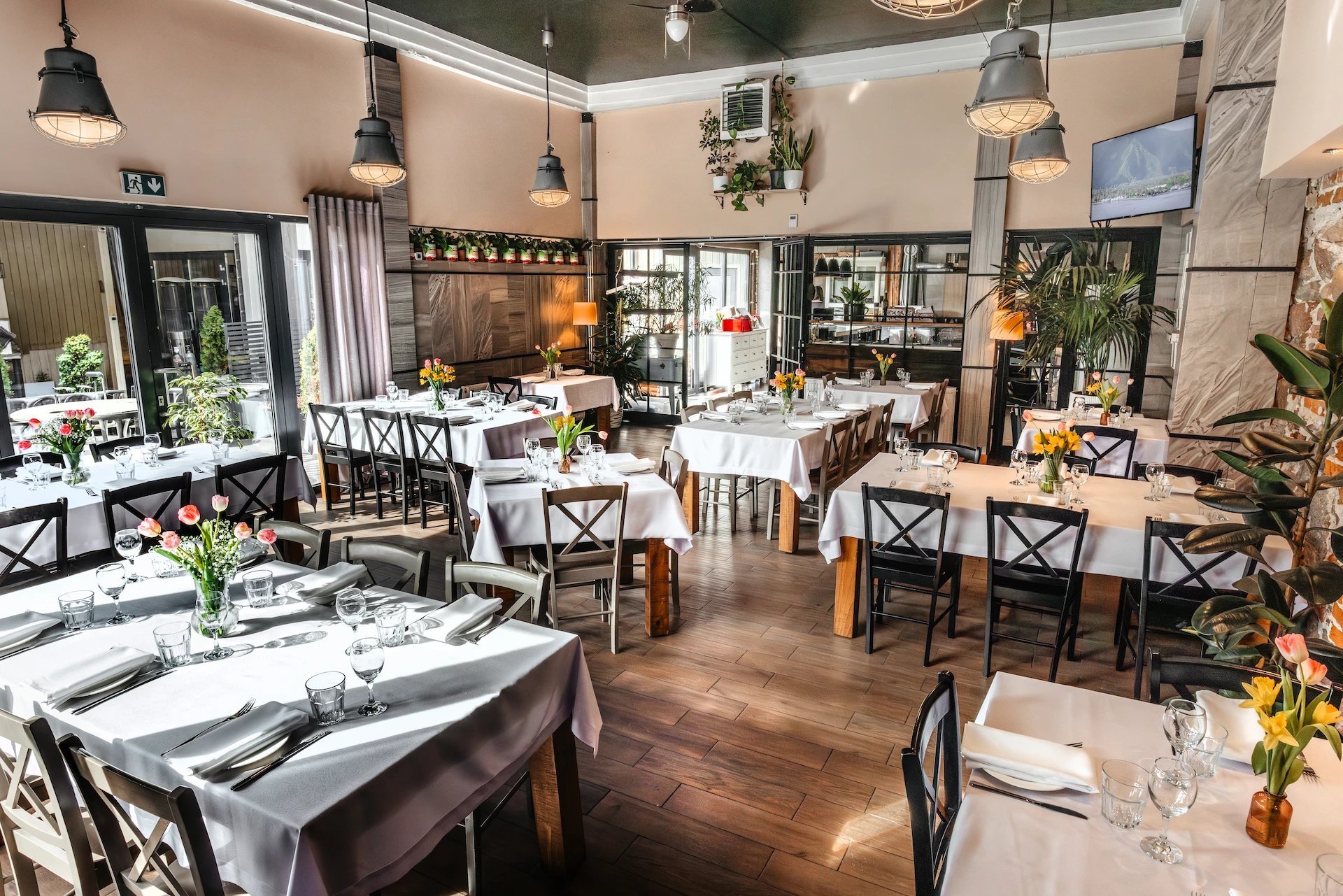 Elegant restaurant dining room with wooden tables, black chairs, white table linens, pendant lighting, and indoor plants throughout the bright space.